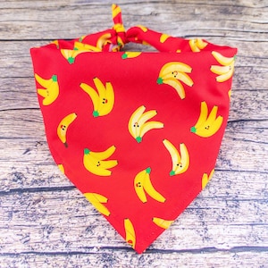 May include: Red bandana with a repeating pattern of yellow bananas. The bananas have green stems and small black dots. The bandana is tied at the top. The background is a wooden surface.
