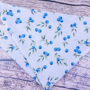 May include: A light blue bandana with a repeating pattern of blue blueberries, green leaves, and small white flowers. The bandana is folded in a triangle shape, with the pattern visible on the fabric.