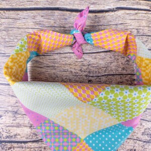 May include: A colorful dog bandana with a patchwork design. The bandana features a variety of patterns in shades of pink, yellow, green, and blue. The top is tied with a pink knot.