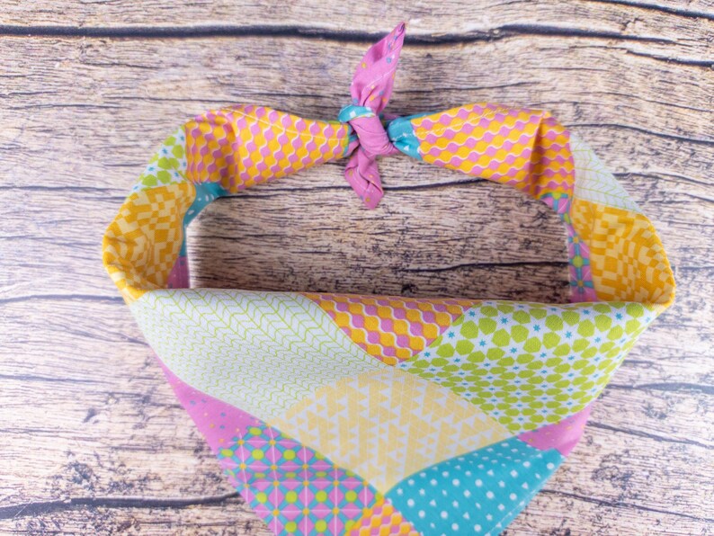 May include: A colorful dog bandana with a patchwork design. The bandana features a variety of patterns in shades of pink, yellow, green, and blue. The top is tied with a pink knot.