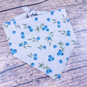 May include: A white pet bandana with a pattern of blue blueberries, green leaves, and small white flowers. The bandana is tied at the top, ready to be worn. The fabric has a soft, slightly textured appearance.
