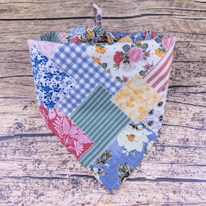 May include: A patchwork dog bandana with a variety of floral, gingham, and striped patterns in shades of blue, pink, yellow, and white. The bandana is tied at the top, ready to be worn.