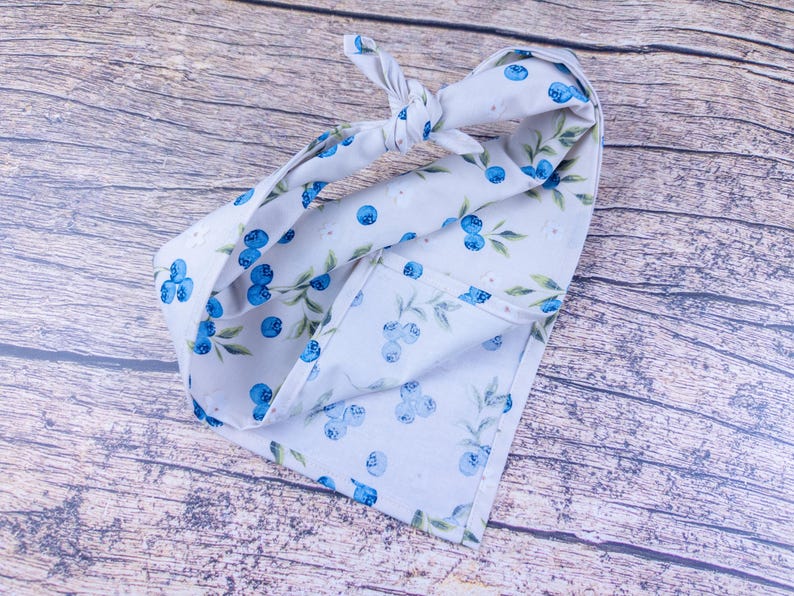 May include: A light blue fabric head wrap with a blueberry and leaf pattern. The head wrap is tied in a knot at the top. The fabric is folded over on itself.