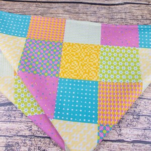 May include: A colorful patchwork bandana featuring squares of various patterns and colors. The design includes pink, yellow, turquoise, and white squares with polka dots, floral, and geometric designs. The bandana is folded in a triangle shape.