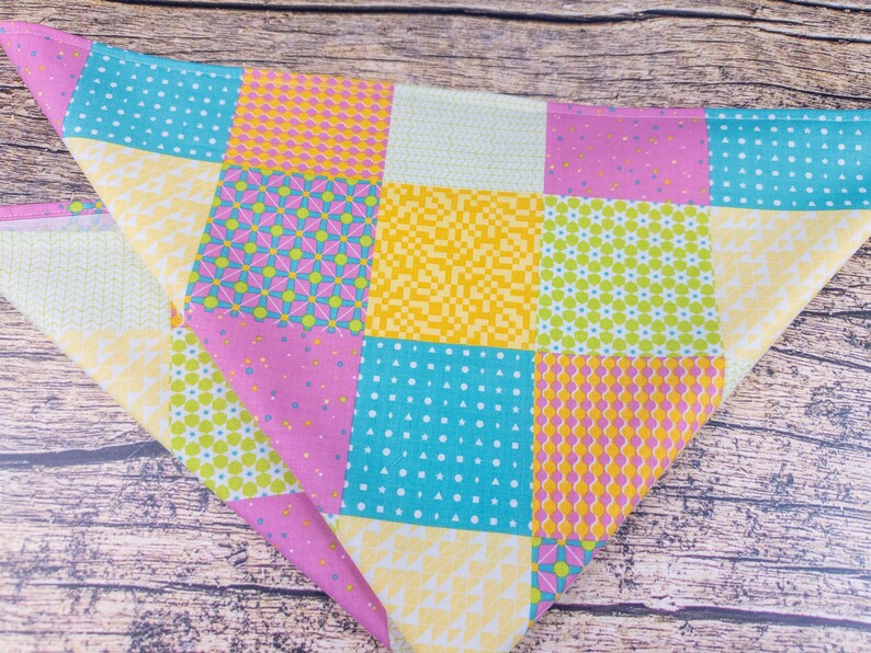 May include: A colorful patchwork bandana featuring squares of various patterns and colors. The design includes pink, yellow, turquoise, and white squares with polka dots, floral, and geometric designs. The bandana is folded in a triangle shape.