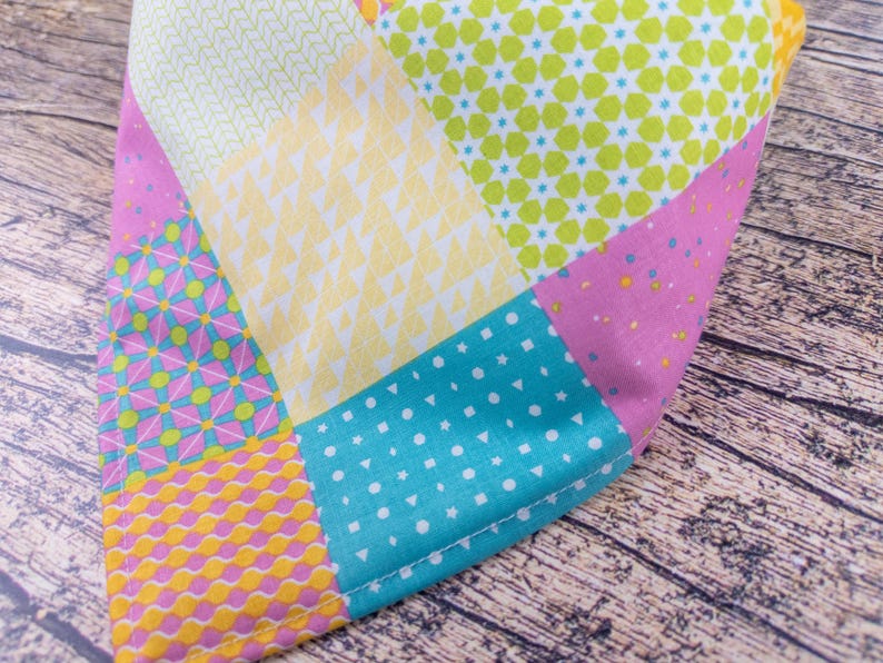 May include: A colorful patchwork fabric with various geometric patterns. The design includes squares and triangles in shades of pink, yellow, turquoise, and green. The fabric is likely cotton or a similar material.