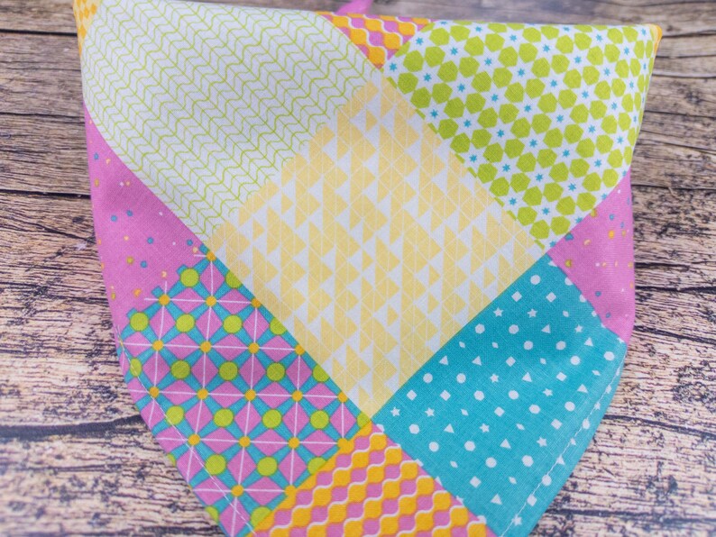 May include: A colorful patchwork bandana featuring various patterns and colors. The bandana is divided into sections of pink, yellow, green, and blue, each with unique designs like polka dots, geometric shapes, and floral patterns. The fabric appears to be cotton.