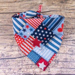 May include: A patriotic dog bandana with a patchwork design featuring red, white, and blue stars and stripes. The bandana is tied at the top with a red and white striped ribbon. The overall theme is Americana.