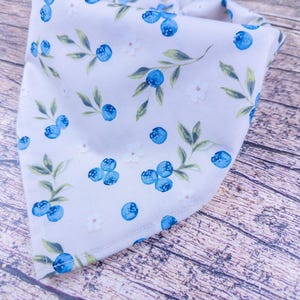 May include: A light gray bandana with a pattern of blue blueberries, green leaves, and small white flowers. The bandana is folded in a triangle shape, with the pattern visible on the front. The background is a wooden surface.