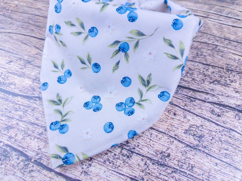 May include: A light gray bandana with a pattern of blue blueberries, green leaves, and small white flowers. The bandana is folded in a triangle shape, with the pattern visible on the front. The background is a wooden surface.
