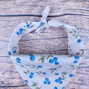 May include: A white pet bandana with a pattern of blue blueberries and green leaves. The bandana is tied at the top, creating a triangle shape. The fabric has a soft, slightly textured appearance.