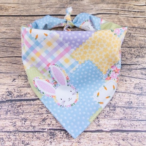May include: A patchwork dog bandana with a bunny face applique. The bandana features a variety of pastel fabrics, including gingham, polka dots, and floral prints. The bunny applique has pink ears and a floral wreath around its face. The bandana is tied at the top.
