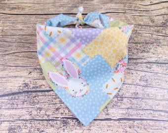 May include: A patchwork dog bandana with a bunny face applique. The bandana features a variety of pastel fabrics, including gingham, polka dots, and floral prints. The bunny applique has pink ears and a floral wreath around its face. The bandana is tied at the top.