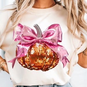 Op de afbeelding: Een wit T-shirt met een gouden en oranje glitterpompoen met een roze strik.