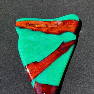 May include: A triangular-shaped resin art piece with a vibrant teal background. Embedded within the resin are pieces of reddish-brown wood, creating a striking contrast. The wood pieces are arranged in a natural, organic pattern, adding visual interest.