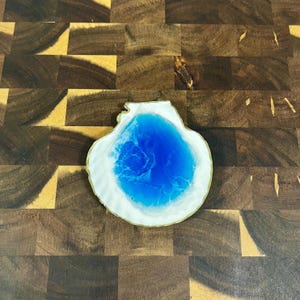 May include: A white and gold resin coaster with a blue resin center. The coaster is shaped like a seashell.