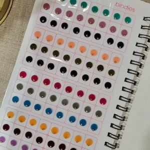 May include: A page from a book showing a variety of colors and styles of round, felt bindies. The bindies are arranged in rows and columns on a white background. The text "bindies" is printed in the upper right corner of the page.