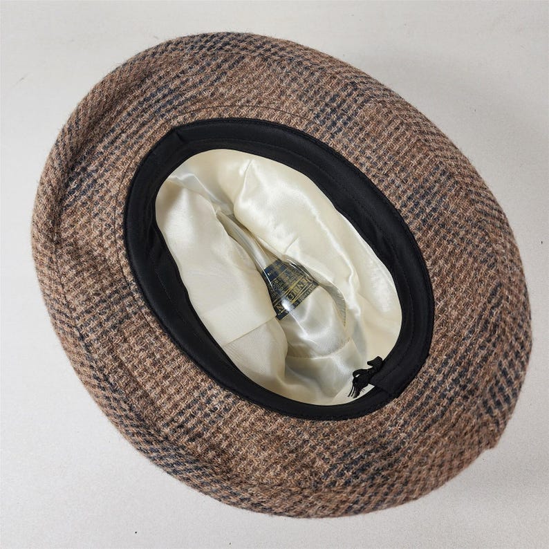 May include: A brown and blue plaid hat with a black brim. The interior is lined with a cream-colored satin fabric. The hat has a classic design, suitable for various occasions.