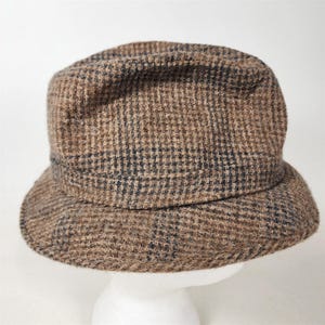 May include: A brown and black plaid hat with a short brim. The hat is made of a textured fabric and has a classic, vintage style. The hat is displayed on a white mannequin head.