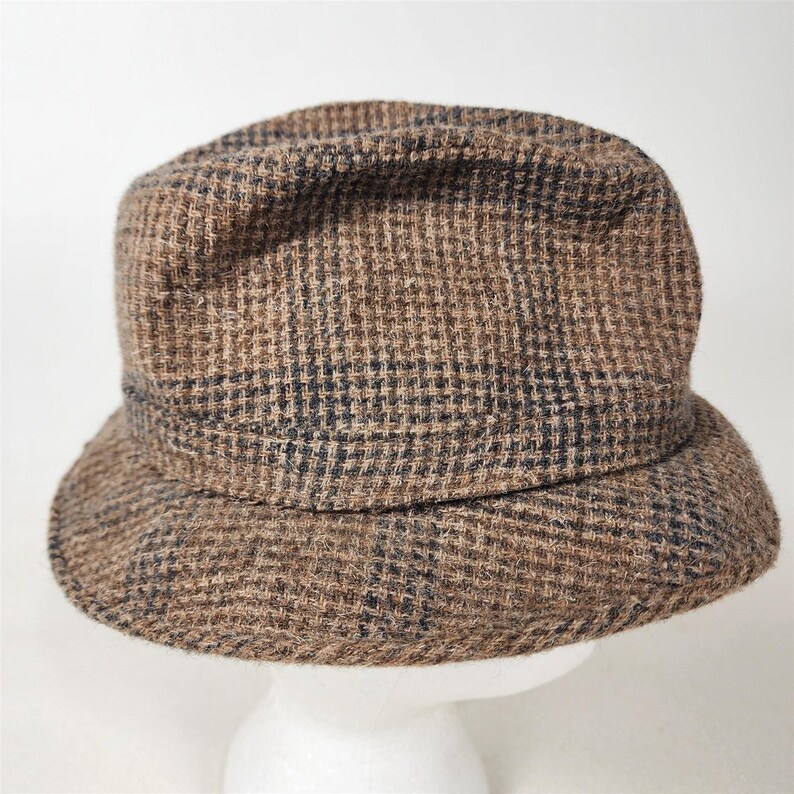 May include: A brown and black plaid hat with a short brim. The hat is made of a textured fabric and has a classic, vintage style. The hat is displayed on a white mannequin head.