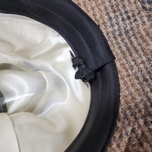 May include: Close-up of a tweed hat's interior. The hat features a brown and black patterned exterior, a black brim, and a cream-colored satin lining. A black drawstring is visible near the brim.