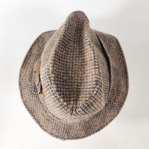 May include: A brown tweed fedora hat with a plaid pattern. The hat features a small feather accent on the side. The hat has a classic, vintage style.