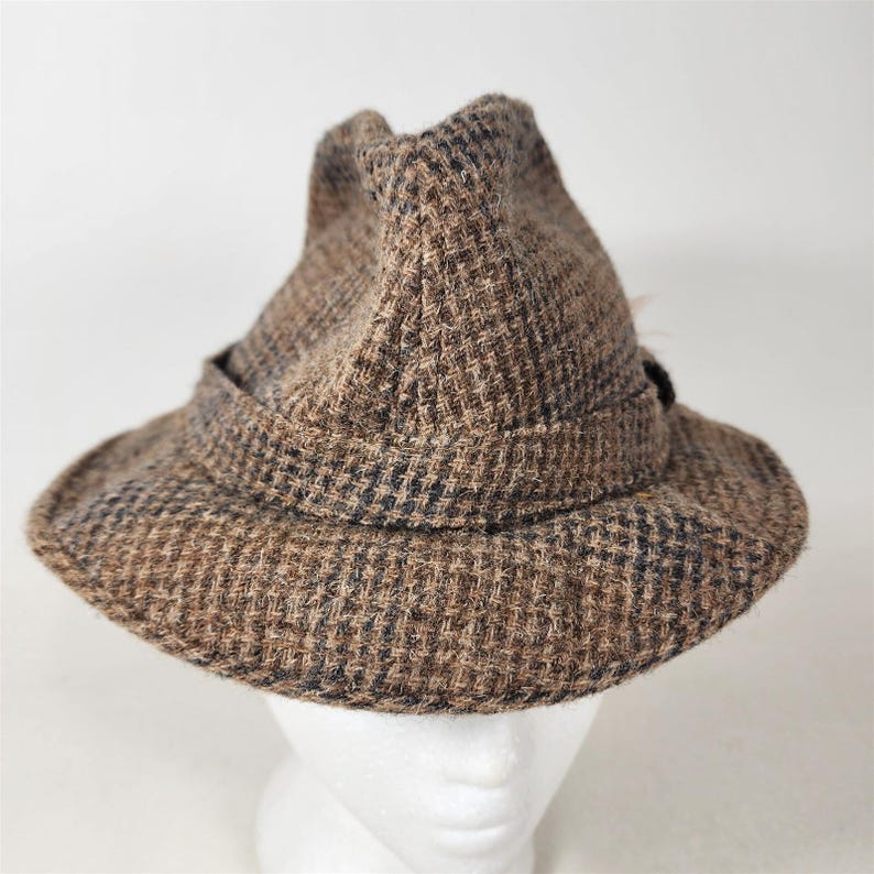 May include: A brown tweed hat with a dark band and a small feather detail. The hat has a classic fedora shape with a slightly upturned brim. The fabric has a woven pattern in shades of brown and black.