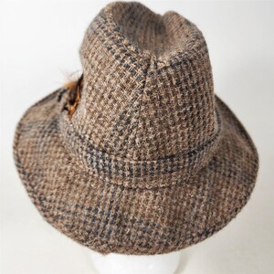 May include: A brown tweed hat with a dark band and a small feather accent. The hat has a classic fedora shape with a slightly indented crown and a brim that curves downward. The tweed fabric has a subtle checkered pattern.