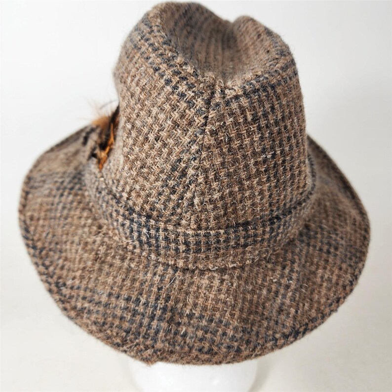 May include: A brown tweed hat with a dark band and a small feather accent. The hat has a classic fedora shape with a slightly indented crown and a brim that curves downward. The tweed fabric has a subtle checkered pattern.