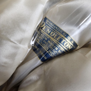 May include: Close-up of a cream-colored fabric with a blue and gold label. The label reads "PENDLETON" and "100% Virgin Wool." The fabric is wrapped in clear plastic.