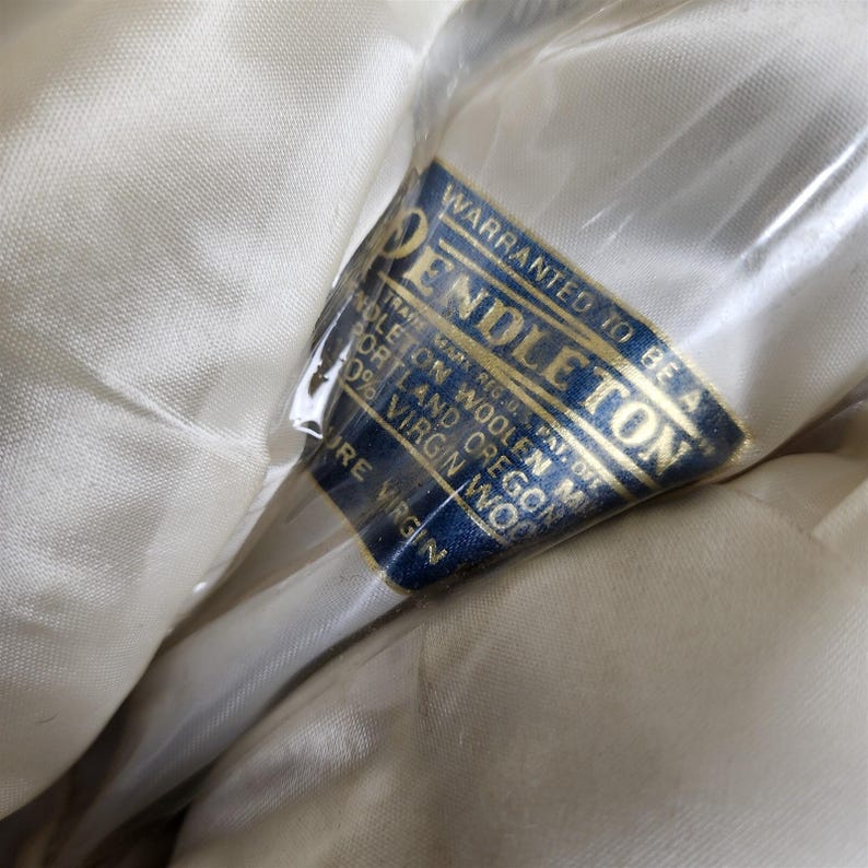 May include: Close-up of a cream-colored fabric with a blue and gold label. The label reads "PENDLETON" and "100% Virgin Wool." The fabric is wrapped in clear plastic.