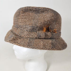 May include: Brown tweed cloche hat with a plaid pattern. The hat features a decorative band and a small feather accent. The hat is displayed on a white mannequin head.