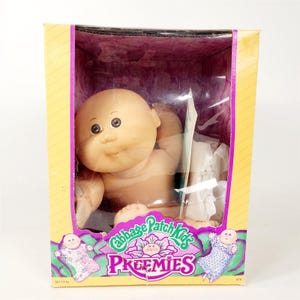 May include: A Cabbage Patch Kids Preemies doll in its original box. The doll has a bald head, round face, and brown eyes. The box is yellow with a purple interior and the Cabbage Patch Kids logo.