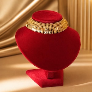 May include: A gold choker necklace featuring a wide, intricate band with a series of small, dangling, leaf-shaped metallic charms along the bottom edge, displayed on a red velvet bust stand to showcase the detailed craftsmanship of the piece.
