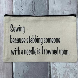 May include: A rectangular beige canvas zipper pouch featuring black text that reads: Sewing because stabbing someone with a needle is frowned upon. The pouch has a black zipper closure along the top edge and a simple, flat construction.