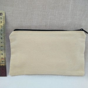 Puede incluir: Una bolsa de lona de color blanco roto con cremallera negra. La bolsa mide aproximadamente 20 cm de largo y 10 cm de ancho.