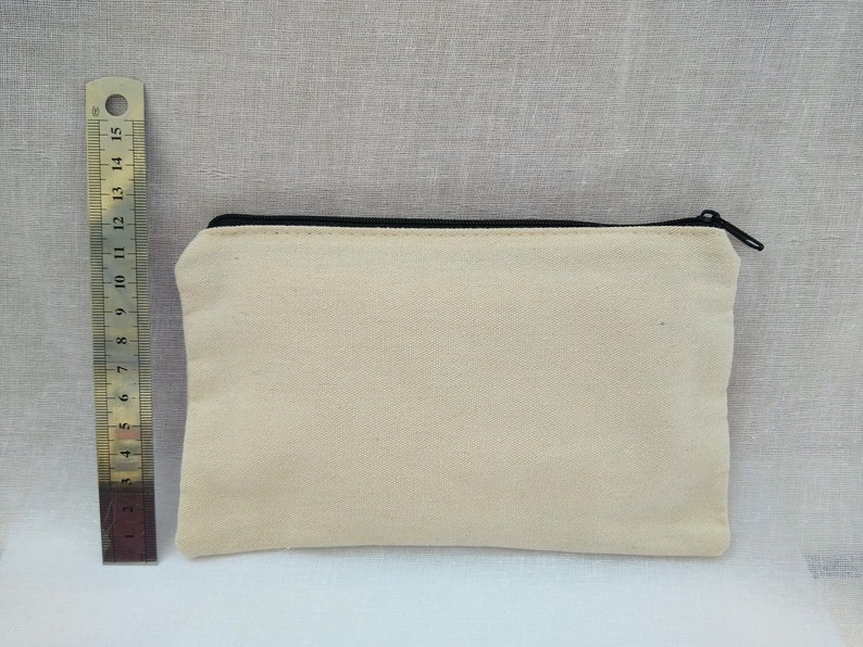 Puede incluir: Una bolsa de lona de color blanco roto con cremallera negra. La bolsa mide aproximadamente 20 cm de largo y 10 cm de ancho.