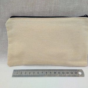 Puede incluir: Una bolsa de lona de color beige claro con cremallera negra. La bolsa est&aacute; colocada sobre una superficie texturizada de color gris claro.