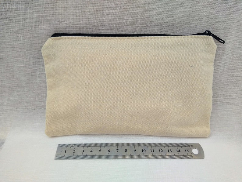Puede incluir: Una bolsa de lona de color beige claro con cremallera negra. La bolsa est&aacute; colocada sobre una superficie texturizada de color gris claro.
