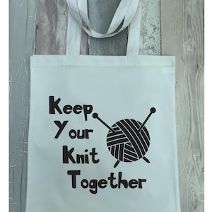May include: White tote bag with black text and a black yarn ball graphic. The text reads "Keep your knit together."