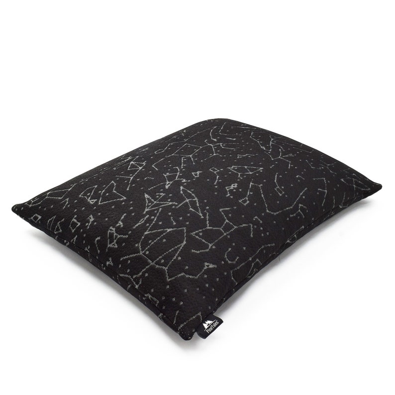 Puede incluir: Una almohada rectangular negra con un patr&oacute;n impreso en blanco de varias constelaciones y estrellas. La tela tiene una apariencia texturizada y una peque&ntilde;a etiqueta de tela negra con el texto blanco PineTales est&aacute; unida a la esquina inferior.