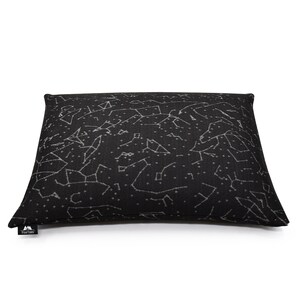Puede incluir: Un coj&iacute;n rectangular negro con un patr&oacute;n celestial blanco de constelaciones y estrellas en toda la superficie. Una peque&ntilde;a etiqueta de tela negra con el texto blanco PineTales est&aacute; unida a la esquina inferior izquierda del coj&iacute;n.
