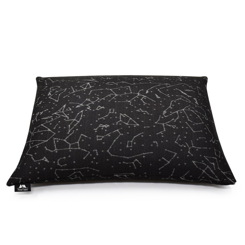 Puede incluir: Un coj&iacute;n rectangular negro con un patr&oacute;n celestial blanco de constelaciones y estrellas en toda la superficie. Una peque&ntilde;a etiqueta de tela negra con el texto blanco PineTales est&aacute; unida a la esquina inferior izquierda del coj&iacute;n.