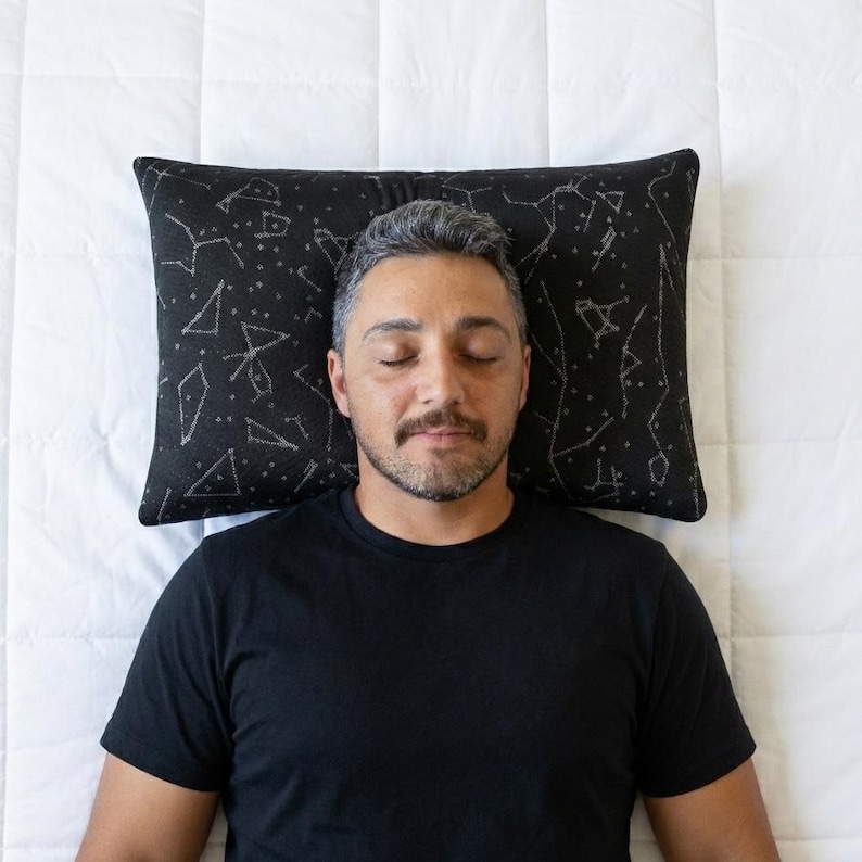 Puede incluir: Una almohada rectangular con una funda de tela negra y un patr&oacute;n estampado en blanco de varias constelaciones y estrellas. La almohada est&aacute; colocada horizontalmente, creando un contraste oscuro sobre la superficie acolchada blanca.