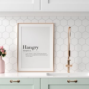 May include: A black and white printable wall art print with the definition of the word "hangry" in a minimalist style. The print is framed in a light wood frame and is hanging on a white wall above a kitchen sink.