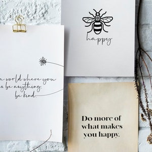 May include: Three inspirational quotes printed on white paper with black ink. The first quote reads "In a world where you can be anything, be kind." The second quote reads "happy" with a bee illustration. The third quote reads "Do more of what makes you happy."