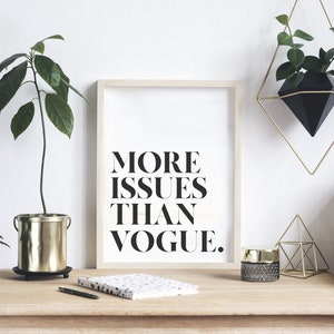 May include: A framed print with the text "MORE ISSUES THAN VOGUE."  The print is on a wooden desk with a gold planter, a notebook, a pen, a gold candle holder, and a gold geometric sculpture.