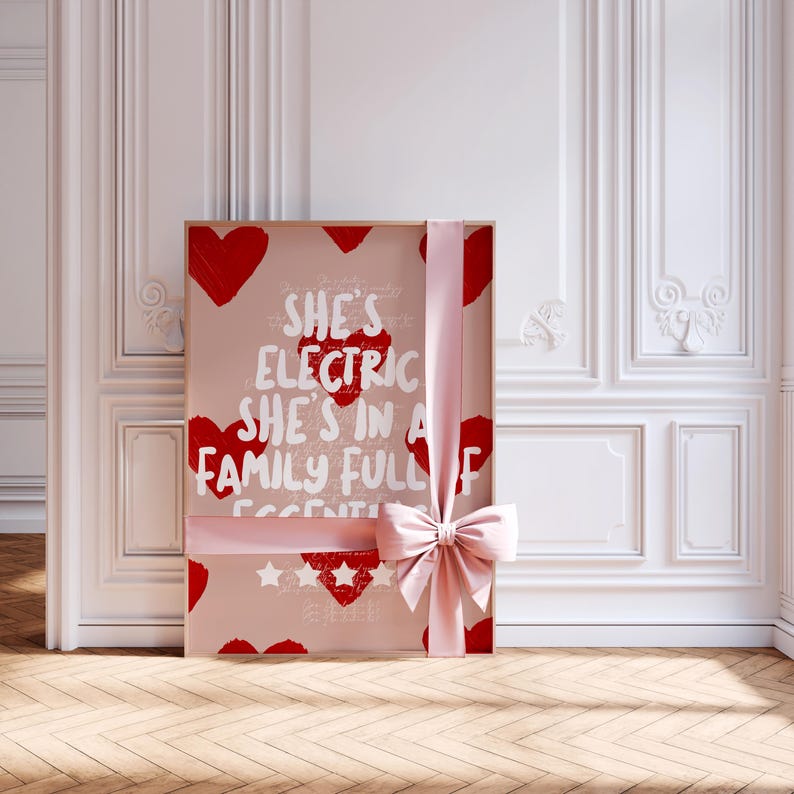 May include: A framed print with a pink background, red hearts, and the text "She's Electric She's in a Family Full of Essentials." A light pink ribbon with a bow wraps around the frame. The print is displayed against a white wall with decorative molding.