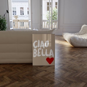 May include: A beige framed print with the text "CIAO BELLA" in white lettering and a red heart in the bottom right corner. The print is leaning against a beige couch in a modern living room.