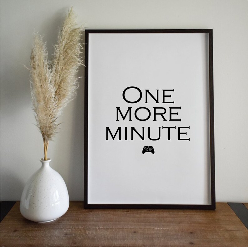 Pode incluir: Uma impress&atilde;o emoldurada com as palavras "ONE MORE MINUTE" em preto, com um &iacute;cone de controle de jogo. Um vaso branco com capim-dos-pampas seco est&aacute; &agrave; esquerda. A moldura &eacute; marrom escuro e o fundo &eacute; branco.
